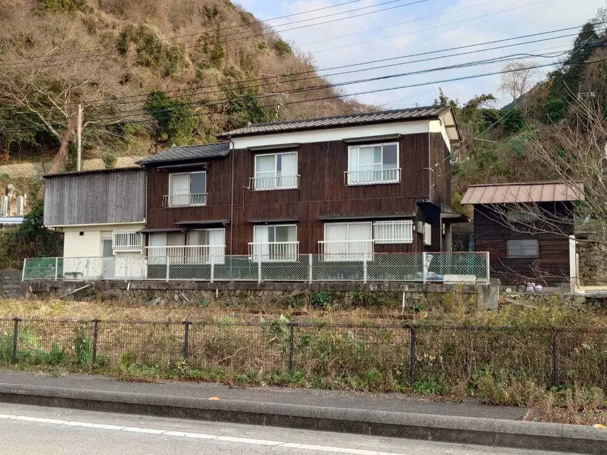 焦げ茶色の板壁が年月を刻む、2階建ての古民家。山を背に、道路を挟んですぐ向こうにJR予讃線が走る