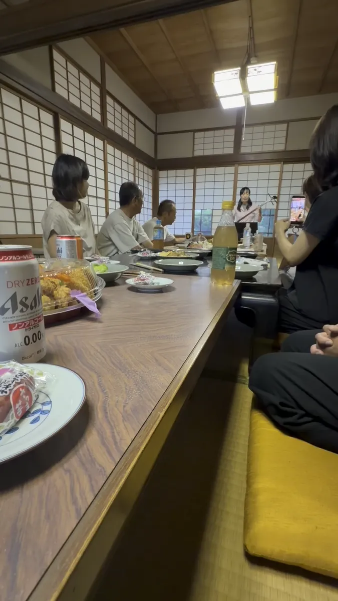 座敷に長机を並べて食事を囲む集まりの様子。奥で三味線を構えた方の演奏がはじまるところ