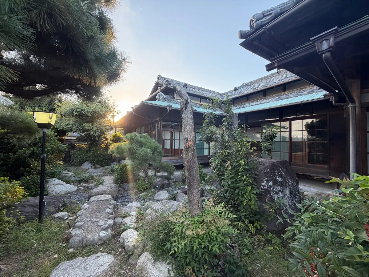松と庭石・飛び石の日本庭園。庭園灯。建物がコの字に庭を囲み、縁側のガラス戸が連なる。松の間から夕日