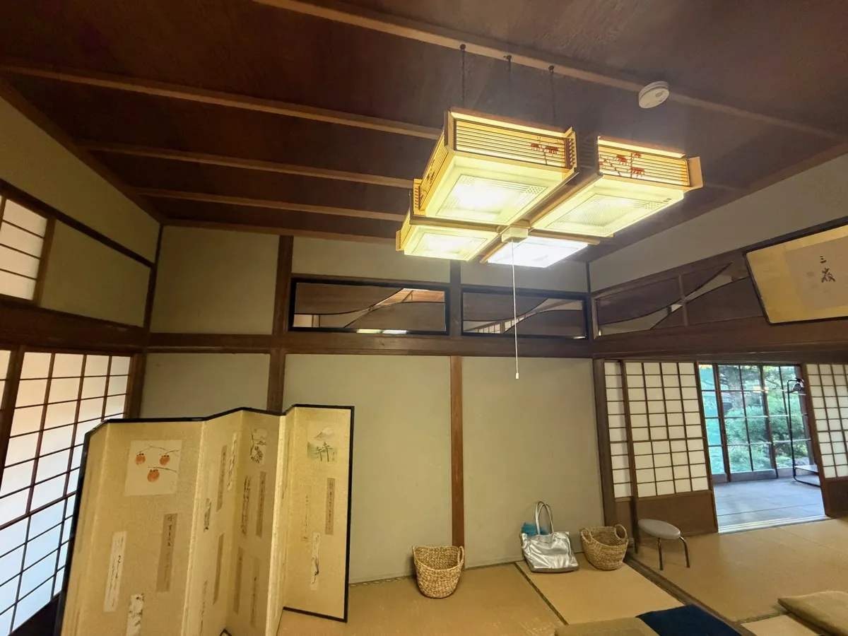 座敷の内部。木の梁と竿縁天井、欄間、障子、和風の吊り下げ照明が2基、絵の入った屏風、畳